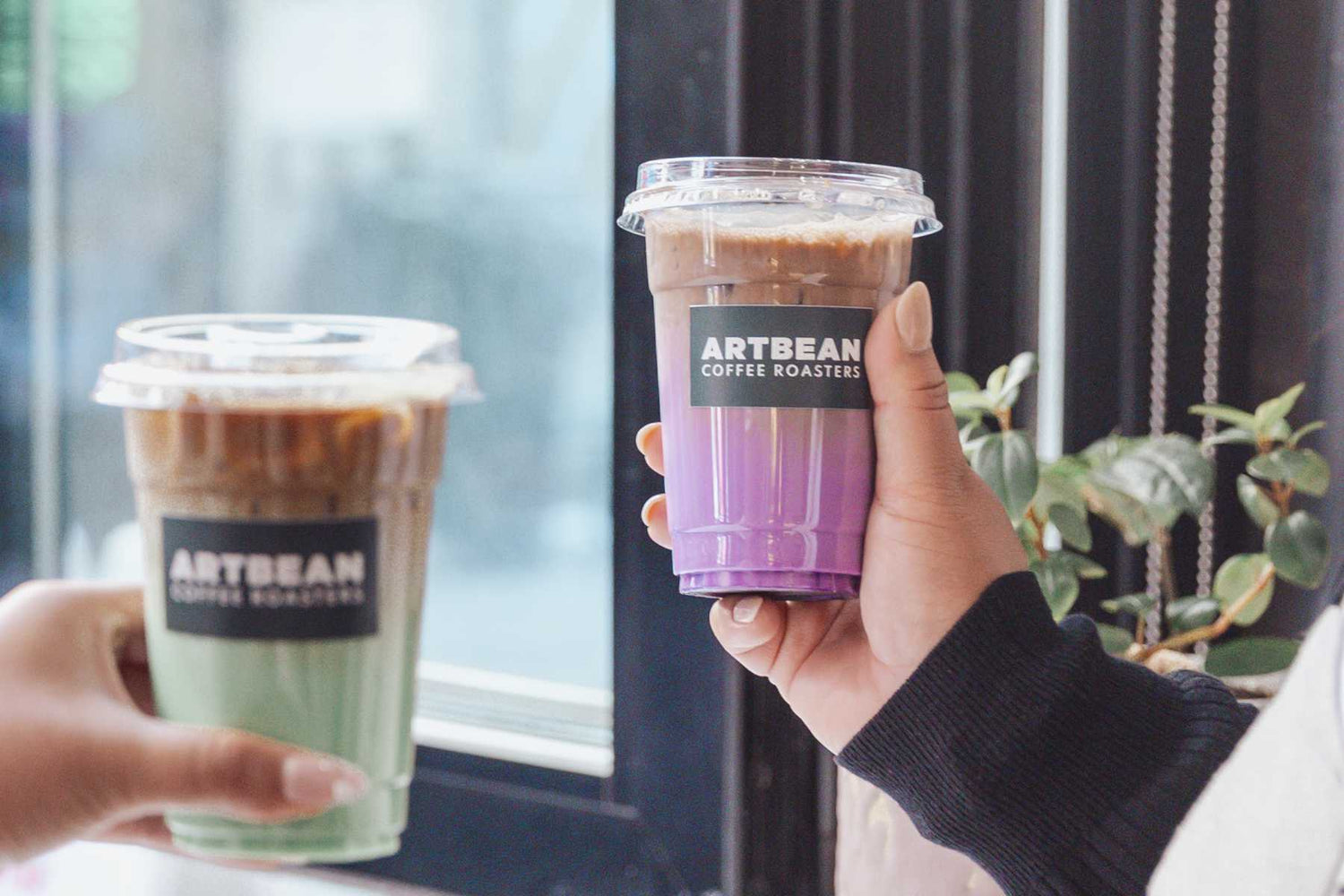 closeup of two hands holding artbean coffee cups. Pandan latte on the left, ube latte on the right